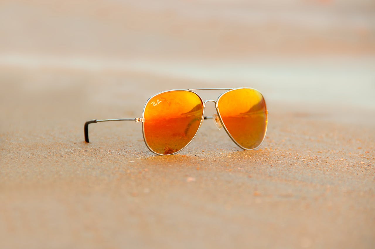 Close-up of aviator sunglasses on sand with orange reflection, evoking a warm summer vibe.